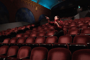Executive Artistic Director, Vanessa Sanchez, gives dancers notes from the audience. Vanessa is seated in the theater, surrounded by empty seats, and is pointing towards the stage.