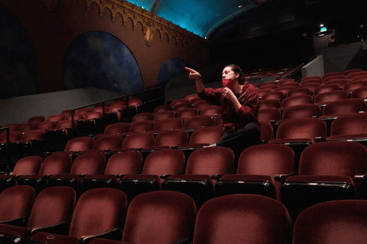 Executive Artistic Director, Vanessa Sanchez, gives dancers notes from the audience. Vanessa is seated in the theater, surrounded by empty seats, and is pointing towards the stage. 