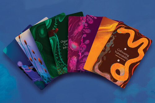 A colorful spread of LANE tarot cards rest on a cloudy dark blue background. Glimpses of illustrations of the natural world, ranging from desert sunsets to tropical vegetation, are visible. The top-most card is fully visible and shows the words 'Cultivating Vulnerability' surrounded by swirling body of a speckled orange snake with sky blue eyes and tongue, shedding a translucent orange and blue skin from its tail. Orange wavy lines radiate from the snake.