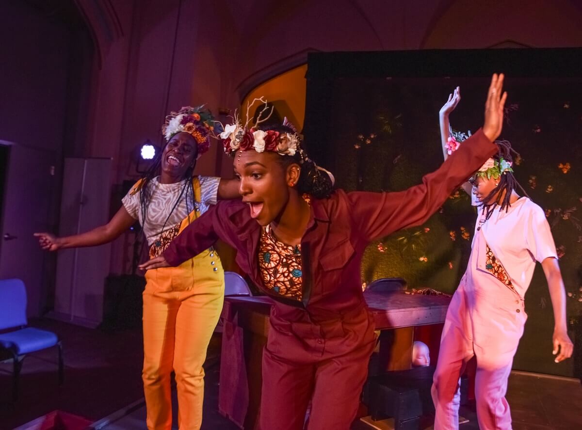 A light brown, slim Black woman, with natural hair pulled back in a low bun, wearing a flower wreath in her hair, in a dark red jumpsuit dances with her hands up in jubilation, behind her a medium brown, Black woman in yellow overalls joins by throwing her arms up and singing loudly " I am deliberate and afraid of nothing", with a flower wreath in her hair. Joining on the right is a light brown woman wearing pink overalls and a white shirt, with medium-length locks, a flower wreath in her hair with hands raised and dancing.