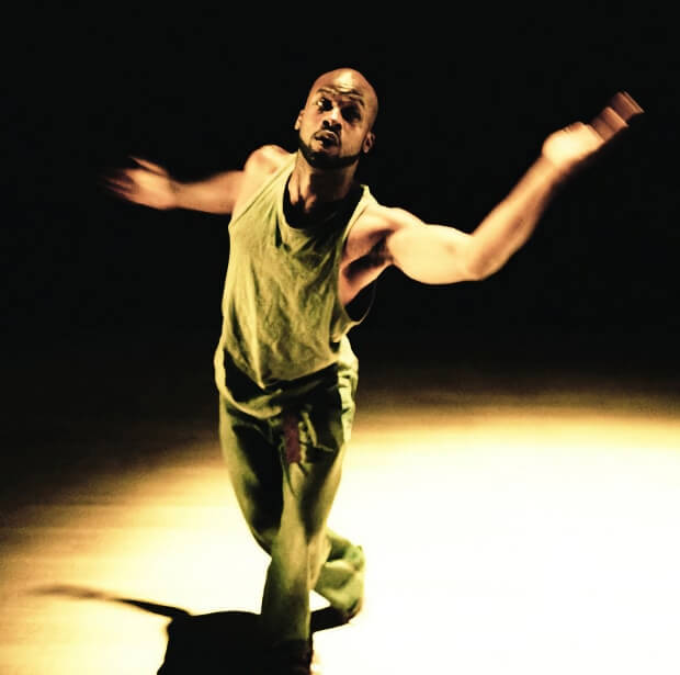 A male dancer in a green tank top and loose green pants performs an expressive contemporary dance move under a warm spotlight on a dark stage. His arms extend outward with motion blur as he leans forward, highlighting the intensity and fluidity of the performance.