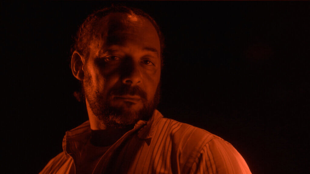 In this still from the film "after angola," a middle-aged bearded man stares somberly at the camera while standing in darkness and bathed in red light.