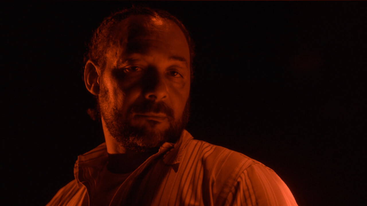 In this still from the film "after angola," a middle-aged bearded man stares somberly at the camera while standing in darkness and bathed in red light.