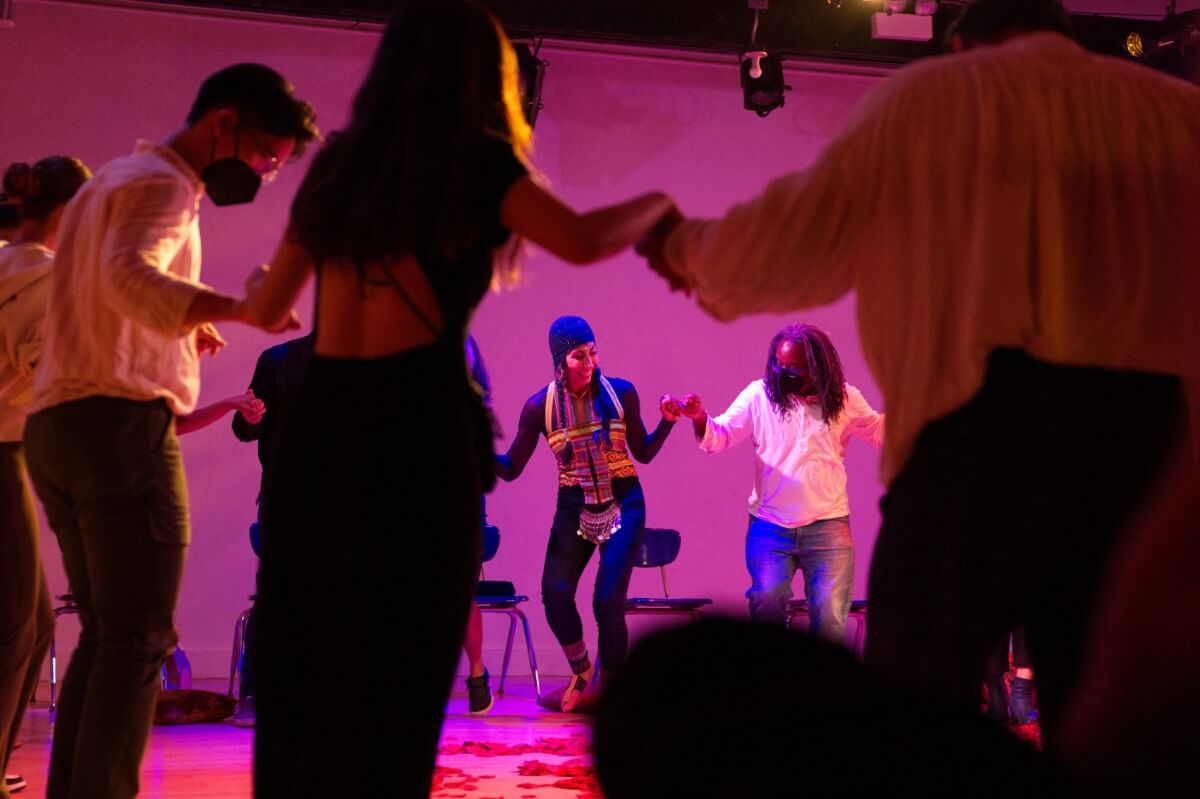 A circle of happy, dancing audience members with performer in blue headpiece and colorful top with tribal prints and blue pants.