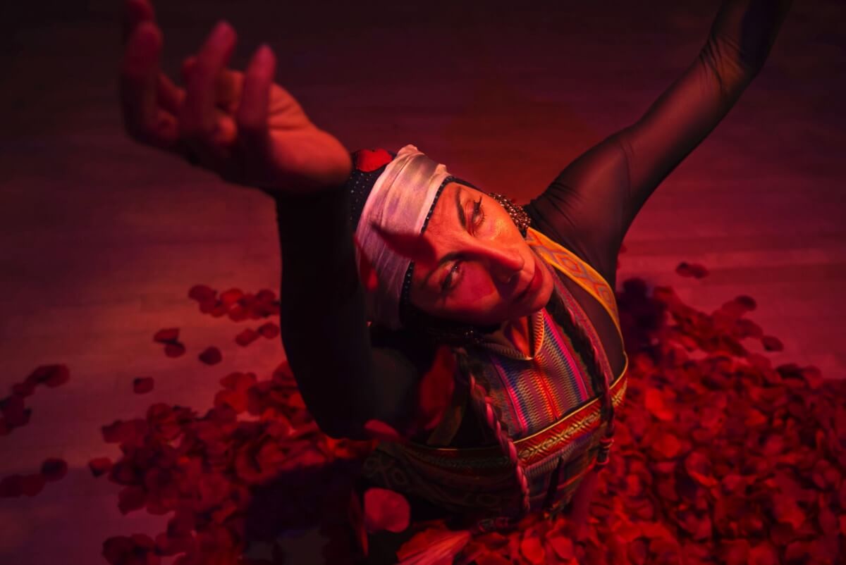 A woman with a blue head piece and white scarf wrapped around her head is kneeling in a pool of red rose petals and she is reaching upwards.