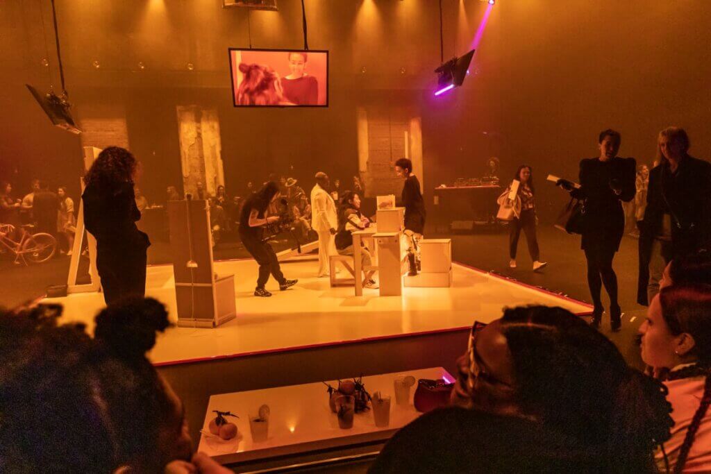 Group of people sitting, standing, and walking around a room that is colorfully lit. There is a square in the middle with audience interaction. Screens suspended in the air show a live feed. People are talking, drinking, and chatting.