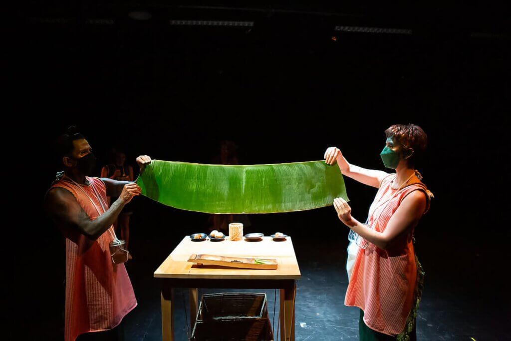 A medium-skinned masked man stands opposite a light-skinned masked woman, with a table between them holding food preparation materials. Both wear aprons and have on face paint. The man holds up one end of a large green banana leaf, while the woman holds the other, stretching it across the table.