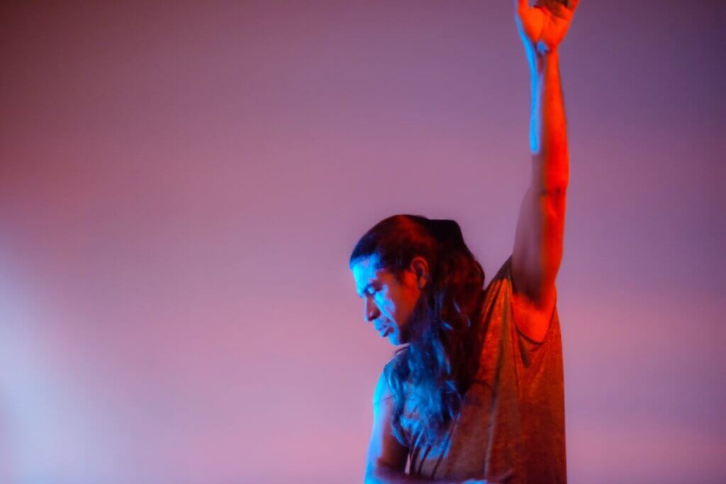 Against a vast, gradient mauve background, Sam Mitchell raises his bare arm high, his hand saturated in warm reddish-orange light. He looks downward with concentration, his profile and long wavy brown hair saturated by a bright blue light. Sam is a lean, middle-aged Indigenous man and wears a sleeveless mottled shirt.