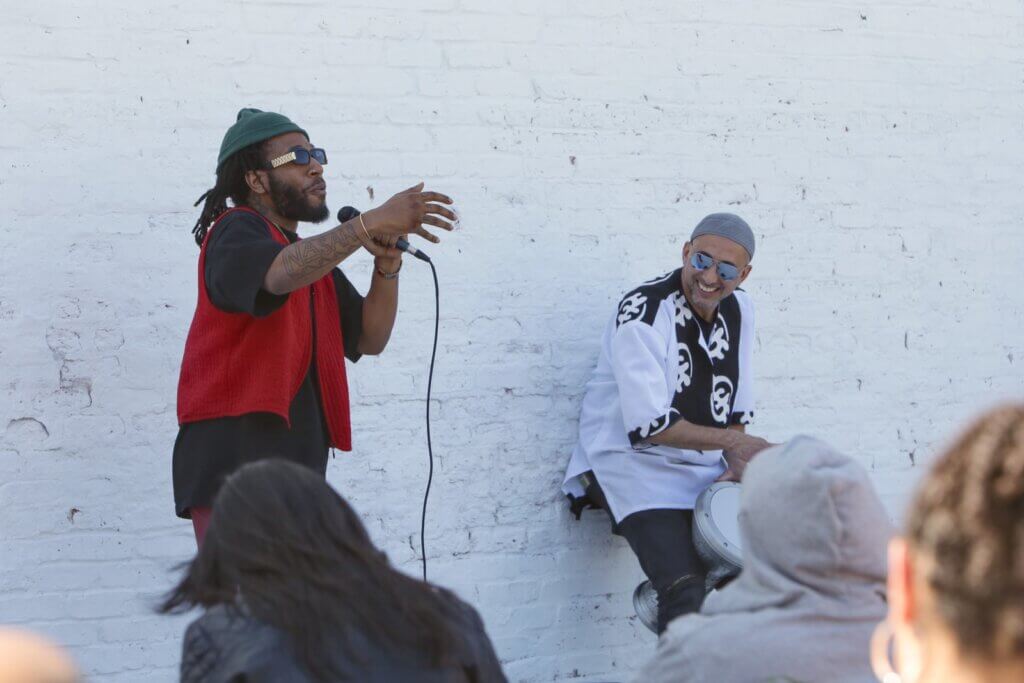 Two men stand in the shadow against a white painted brick wall and address an audience that is only partially visible in the extreme foreground of the composition. The man on the left is a Black bearded man with short dreadlocks that are tied back. He is wearing a black shirt with a knitted red vest, a green knit cap, and black and gold sunglasses. He is holding a microphone and gesturing with his free hand as he speaks. The other man appears to be of Middle Eastern descent. He is wearing a loose-fitting white and black shirt, a grey cap, and mirrored sunglasses, and he is playing a drum held between his knees as he leans back against the brick wall and smiles at the other man.