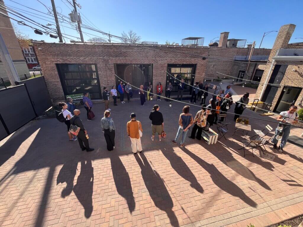 An arial shot of a circular arrangement of people standing in partial shadow in a small red brick courtyard in the afternoon. They appear to be engaged in a group activity and are looking at someone on the right side of the circle.
