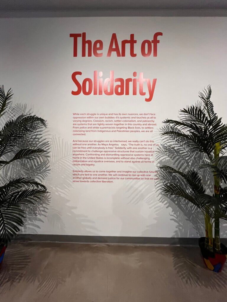 Red text printed directly onto a white wall in an undisclosed location. There is a large title in letters that are 12 to 18 inches tall, followed by three paragraphs of smaller text. The title reads "The Art of Solidarity." The smaller text is unreadable. To the left and right of the text stand potted plants.