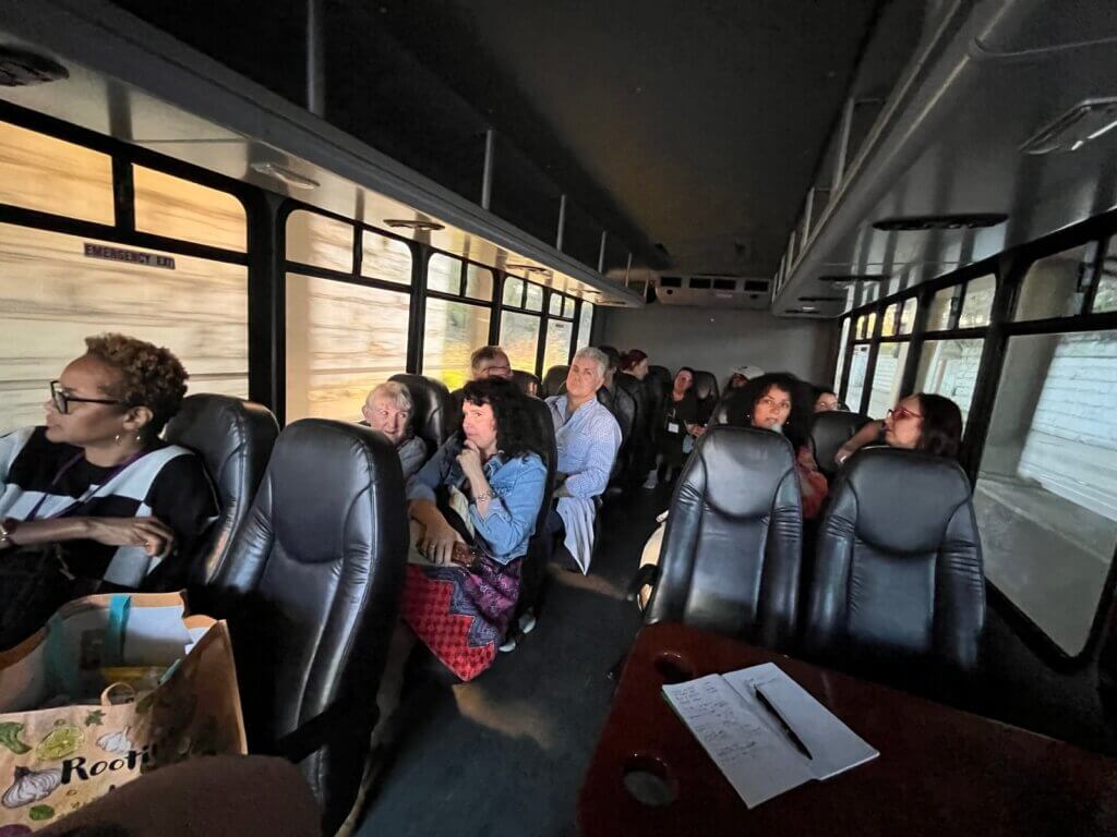 The interior of a bus, with most seats filled with people of various skin tones and genders. On the left side of the photo, a nearby wall blurred by the motion of the bus is visible through the windows. On the right side, a more distant wall can be seen.