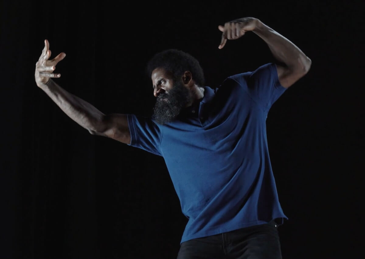 A Black man stands in a dark blue polo shirt mid-lunge with his arms raised and his hands gesturing energetically. His face is turned profile and tilted down so that his thick beard melts into his shirt and the stage light gleams bright against the dark skin of his forehead. His expression is tense, with knit brows, a scrunched nose and a thin mouth.