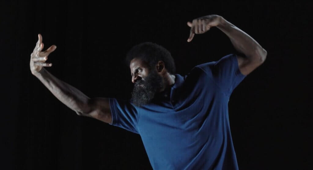 A Black man stands in a dark blue polo shirt mid-lunge with his arms raised and his hands gesturing energetically. His face is turned profile and tilted down so that his thick beard melts into his shirt and the stage light gleams bright against the dark skin of his forehead. James’ expression is tense, with knit brows, a scrunched nose and a thin mouth.