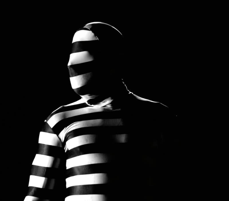 Portrait of a person in a striped black and white full body suit that, in places, blends with the black background. The suit covers his face, which is turned profile. The lighting illuminates half of his body and leaves the other half in dramatic shadow.