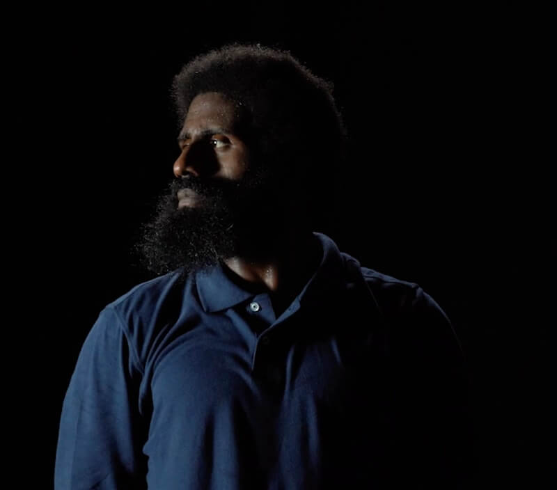 Portrait of a Black man in a dark blue polo. His face is turned profile so that his thick beard catches the light and frames the slope of his shoulder. Half of his body is covered in shadow.