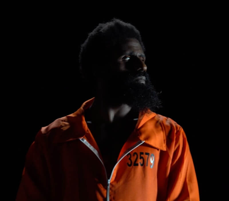 Portrait of a Black man in a zippered orange prison jumpsuit. His face is turned profile, and his hair and beard fade almost completely into the black background. The stage light reflects the white of his eyes—highlighting his downturned gaze, which appears to be hauntingly directed at the number printed on the left breast of the jumpsuit: 32579.