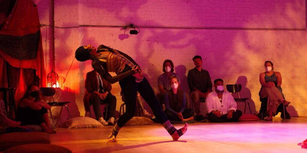 Woman with blue head piece and colorful top with tribal prints and blue pants leaning backwards in front of a circle of seated audience members. The space is decorated and lit in purple colors.
