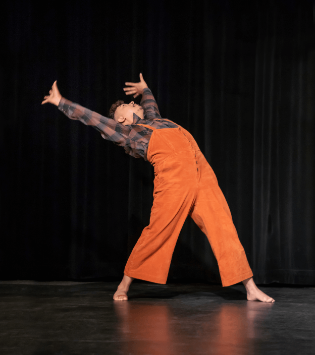 A medium brown skinned Mexican American dancer, wearing an orange overalls with buttons and a brown and black flannel shirt underneath, looks upwards while extending arms up and backwards with arch in back legs in a wide stance. He stands alone on stage with back curtains behind him. The background has a black curtain and a dark floor.