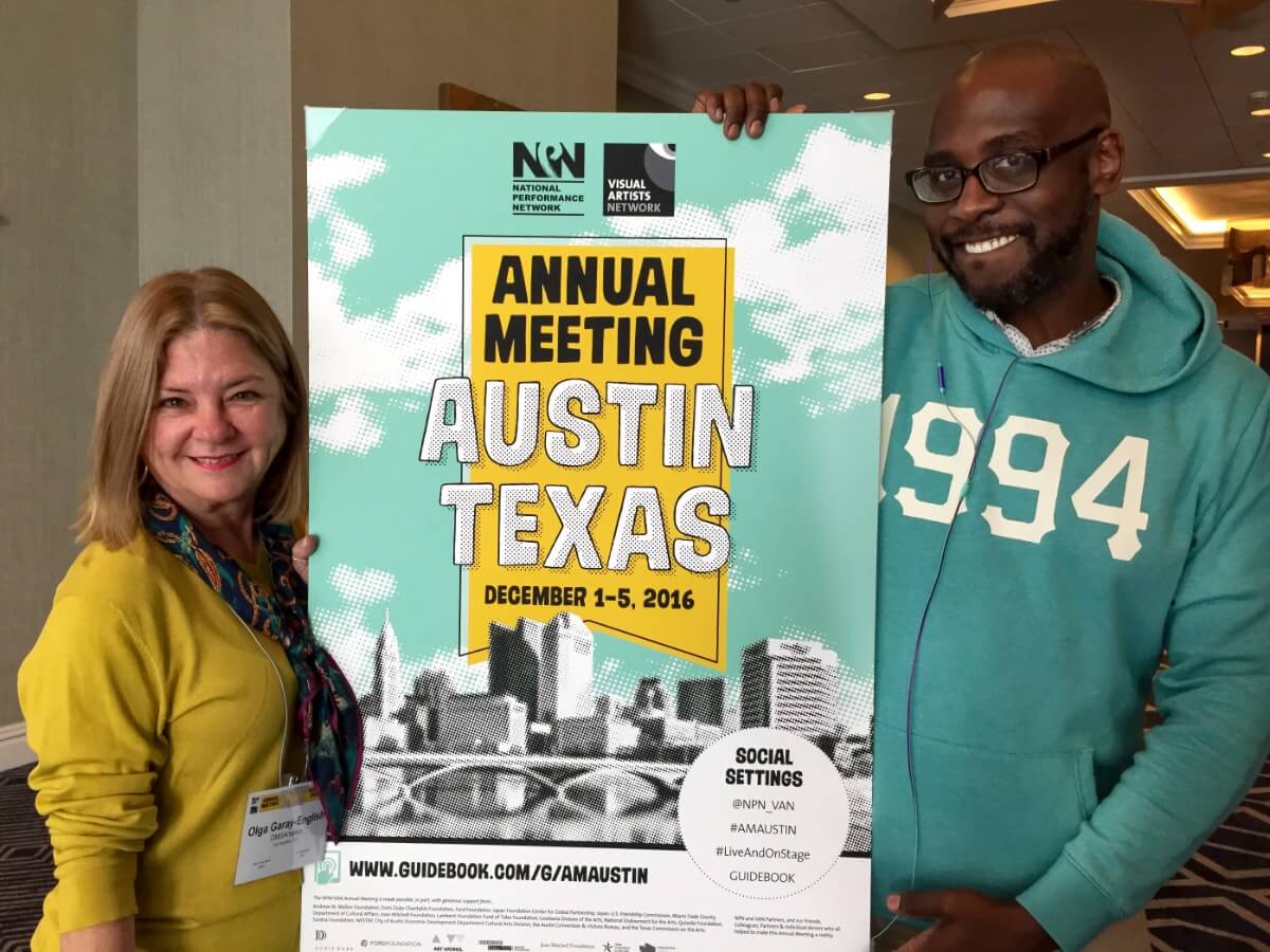 Two people pose smiling on either side of a poster for NPN's 2016 Annual Meeting in Austin, TX. On the left is a light skinned woman with shoulder length blonde hair. She is wearing a yellow shirt with sleeves pushed up just below the elbows, and a tied kerchief that's navy blue with elements of gold, green, and magenta. She has a large name tag hanging from a lanyard around her neck. On the right is a tall, dark skinned man with a bald head and closely cropped short beard. He is wearing glasses and a teal hoodie pullover with the numbers "1994" in white across the chest, and there are headphone wires extending up the front and disappearing into the neck opening of the pullover. The poster is white and teal, with a black and white image of the Austin skyline across the bottom third. The center of the poster is filled with a large yellow ribbon shape outlined in offset black lines, over which are the words "Annual Meeting Austin Texas December 1-5, 2016." Black versions of the logos for NPN and the former Visual Artists Network are placed above the ribbon shape.