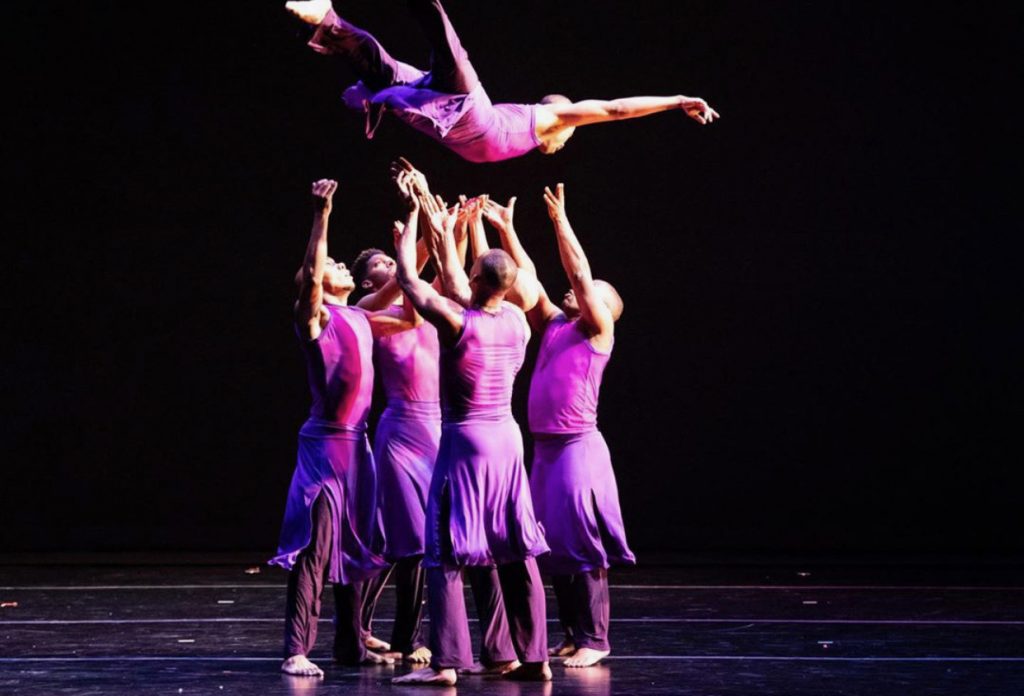 Four dancers in purple on a stage stand with arms extended upward beneath another dancer who is in the air
