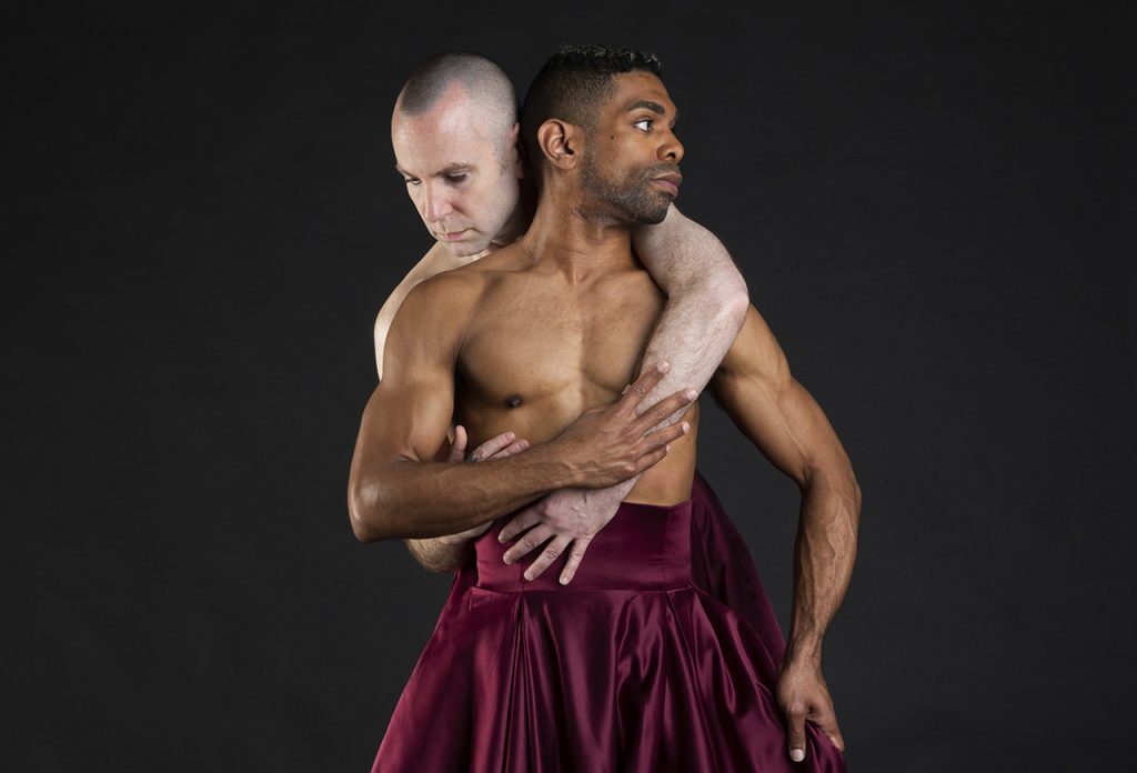 One dancer embraces another from behind. They gaze in opposite directions and both wear maroon velvet skirts.