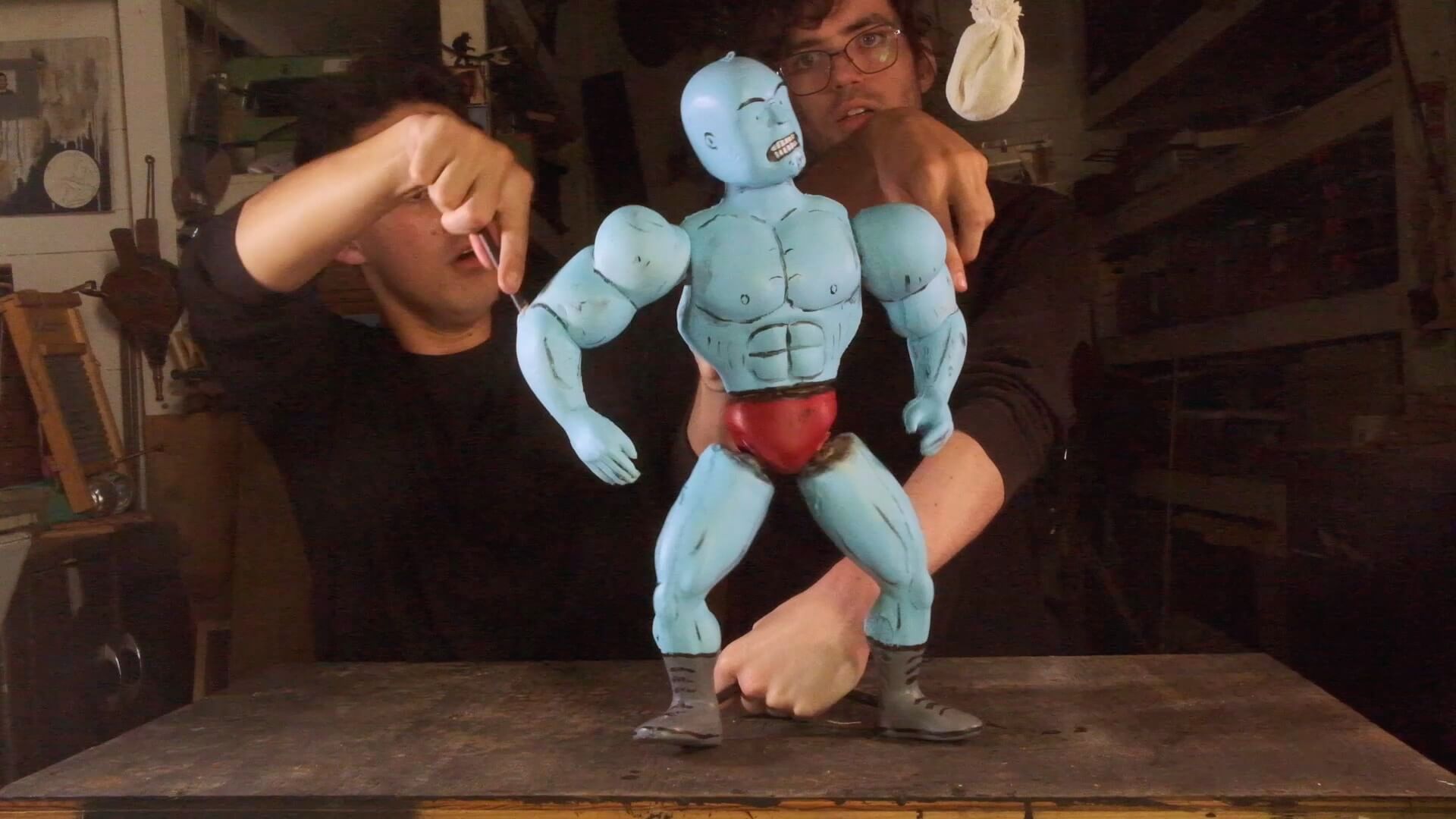 Two young men dressed in black use metal rods and work together to pose a 15-inch-tall foam puppet that looks like a bald, muscular, blue-skinned bodybuilder or professional wrestler, wearing red briefs and black boots. The puppet is scowling and staring up at a miniature white slip bag, or punching bag, used by boxers for training.