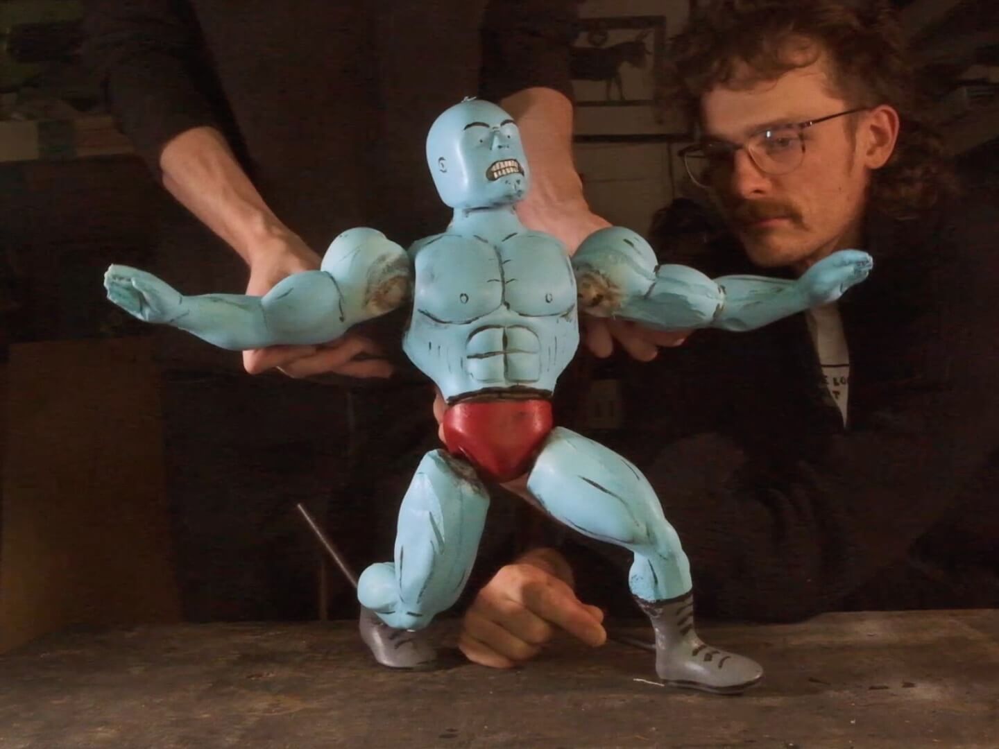 Two young men dressed in black use metal rods and work together to pose a 15-inch-tall foam puppet that looks like a bald, muscular, blue-skinned bodybuilder or professional wrestler, wearing red briefs and black boots. The puppet is scowling and has one leg extended behind him, as if he's lunging forward, with his arms outstretched.