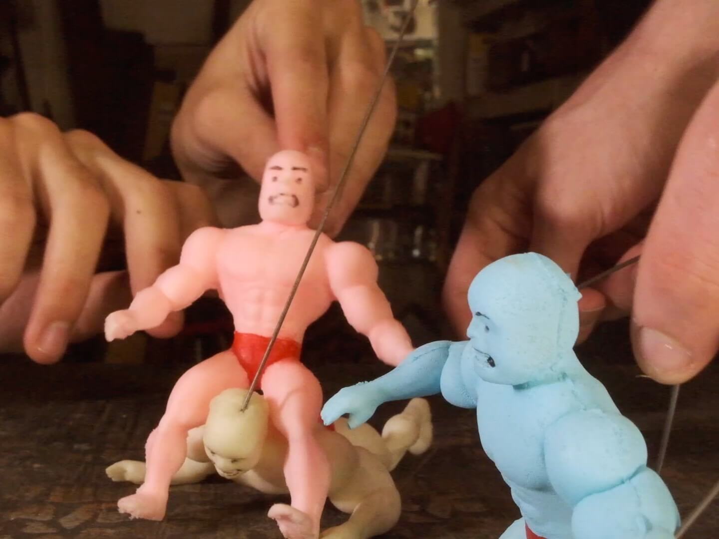 A close-up of two sets of hands using metal rods to pose a trio of 4-inch-tall foam puppets that each look like bodybuilders or professional wrestlers. Each puppet is a single pastel color, bald, barefoot, and wearing red briefs. The yellow puppet is lying with his chest on the ground and lifting his head up, while the pink puppet is straddling the yellow puppet's neck in what looks like a wrestler move. In the foreground, the blue puppet appears to be reacting with alarm to the struggle between the yellow and pink puppets.