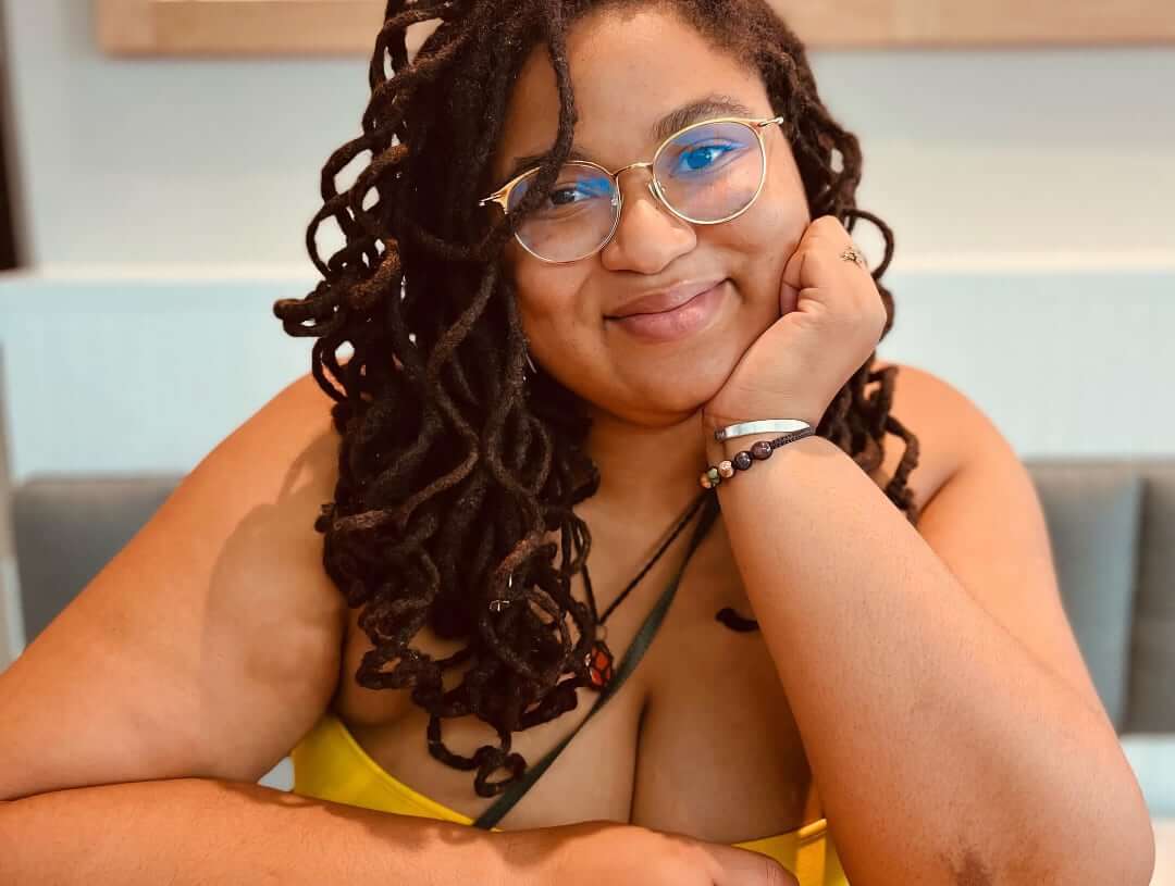 A medium-skinned woman with locs and big glasses rests her chin on one hand and smiles.