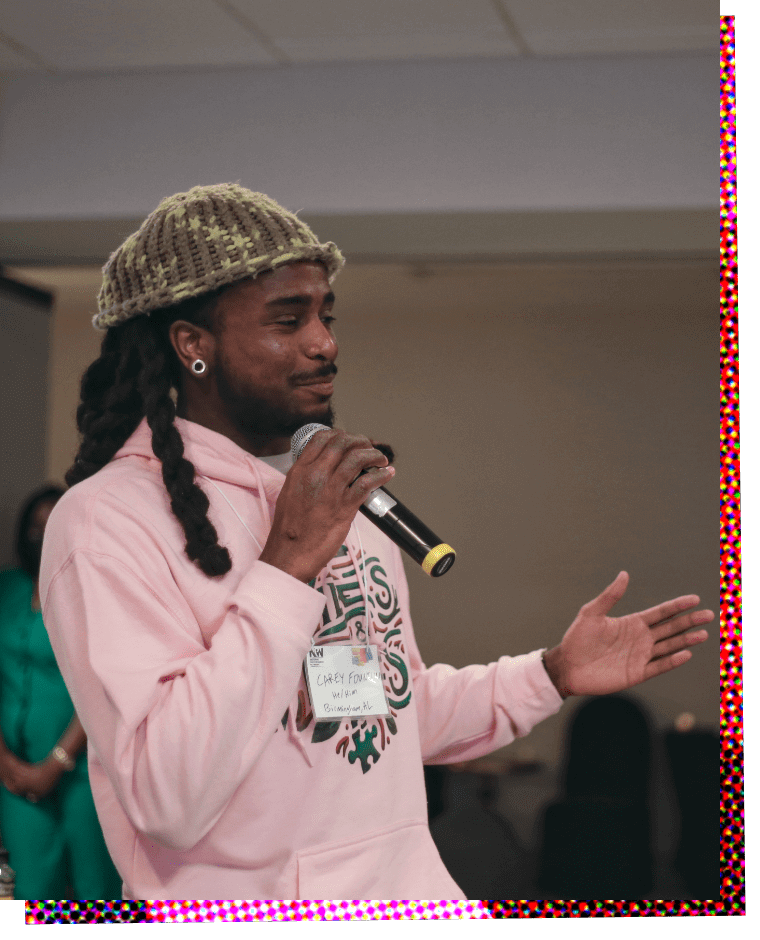 Photograph of Carey Fountain, a Black man dreadlocks pulled back and wearing a greyish-green fuzzy knit cap. He is standing in three-quarter profile facing left, and holds a microphone in his right hand as he gestures with his right hand. He is wearing a light pink cotton hoodie with a decorative green and maroon pattern on the chest. 