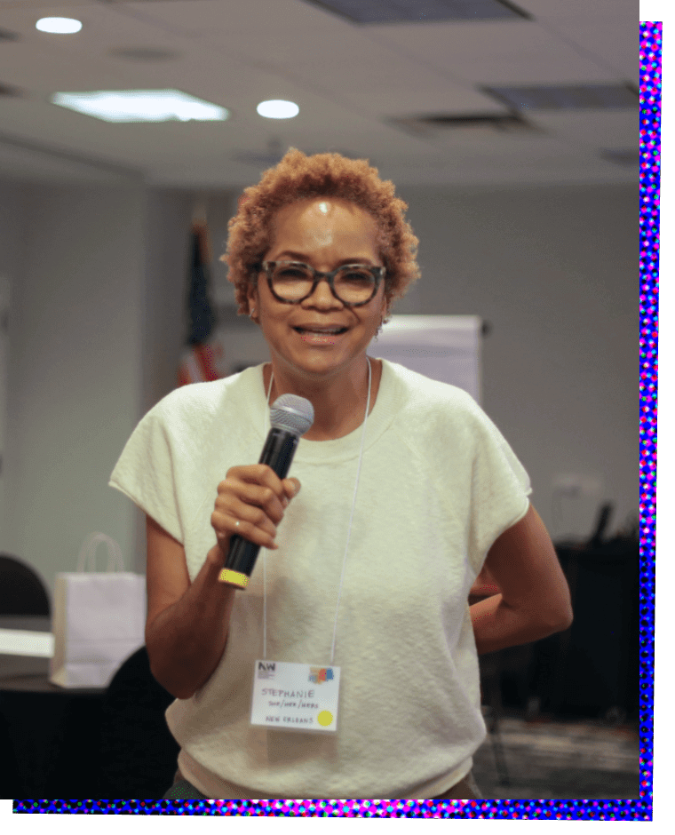 Photograph of Stephanie Atkins, a Black woman. She is standing and facing the camera and smiling as she speaks into a microphone that she holds in her right hand. Her left arm is folded behind her so that only the elbow is visible. She has short red hair and wears black glasses. She is wearing a short sleeved, loosely fitting white t-shirt.