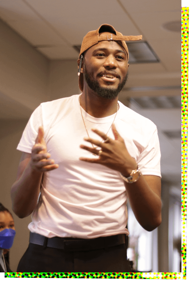 Photograph of Devonta Ravizee, a young Black man with a trim beard, who is standing and gesturing with his hands as he speaks. He is wearing a brown or dark orange baseball cap turned backwards, a light t-shirt that appears to be white or peach colored, and black pants. He has a gold watch on his right wrist.