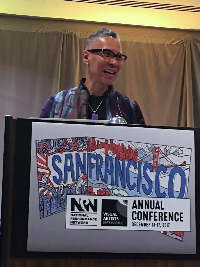 A Black woman with short black and gray hair that's shaved on the sides stands behind a podium. She is wearing black glasses and silver pendant style earrings, a black shirt, a silver necklace from which hangs a large purple stone, and a purple, light blue, and gold patterned jacket. She is smiling as she addresses the unseen audience. Hanging over the front of the podium is a large sign with hand-drawn lettering that reads "San Francisco" over a white, red, and blue mural of some of the city's most recognizable landmarks and imagery, including rows of narrow houses, the Golden Gate Bridge, and the San Francisco Bay. Overlaid across the bottom of the image is a white rectangle that displays black versions of the logos for NPN and the former Visual Artists Network, as well as the words "Annual Conference December 14-17, 2017."