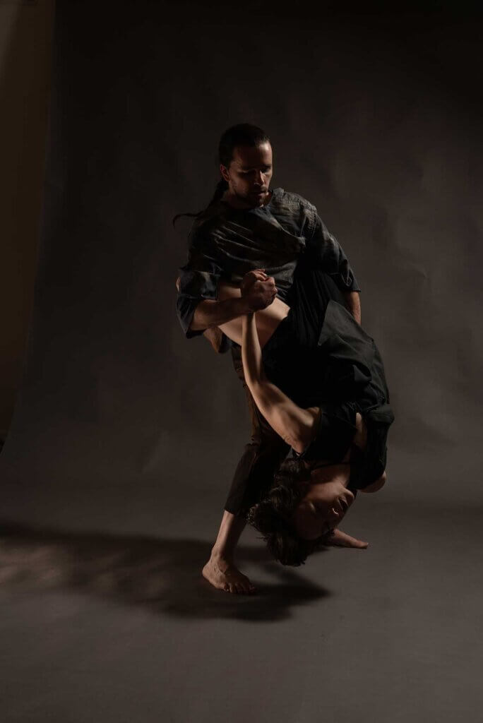 Two white folks (Anna Thompson and Taylor Knight) are partnering in a dimly lit room with a gray background. Anna hangs upside down; their dark hair is wildly suspended in time. They have their legs wrapped around Taylor’s neck and are holding their hand to provide support. Taylor has their hair slicked back into a ponytail braid; they are looking down towards Anna with their left hand providing support on their back.