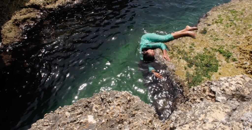 The image looks down on this scene from above: a Palestinian woman amongst waves and stone. She has brown hair and skin and is shaping her body against the curve of a stone, hands open and pressed on the surface covered with green yellow algae. She is wet and also half immersed in the surrounding, rippling blue-green water. Her dress is turquoise, eyes closed, face turning away.