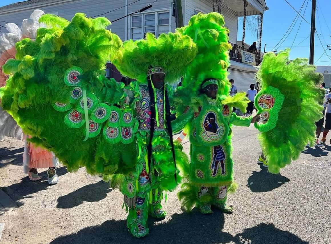 Two Masking Indians pose with their arms around each other in a sunny, crowded street. The camera is positioned to their right. The Masker to the left has his right arm wide-open in a green-feathered multicolor rhinestoned suit. His wing is spread under his feathered arm. He wears a round feathered sun hat on his head with a multi-green feathered war skirt around his waist, and his two braids run almost to the floor. The Masker to the right has her left arm wide-open in a similarly green feathered multicolor rhinestoned suit. She holds a feathered fan in her left hand. A towering, feathered headpiece sits on her head and a fuzzy green-patched skirt around her waist.