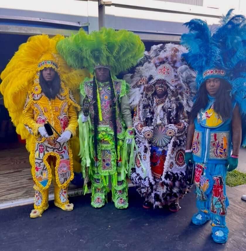 Four Masking Indians pose on the sidewalk with their arms by their sides in different-colored suits. From the left to the right, there is a Masking Indian in a yellow suit, one in a green suit, one in a brown and white suit, and one in a blue suit. All of these suits are decorated in colorful patches, all are beaded except the green one which is rhinestoned. The Masker in the green suit wears a green round feathered sun hat. The others wear large feathered open-face crowns.