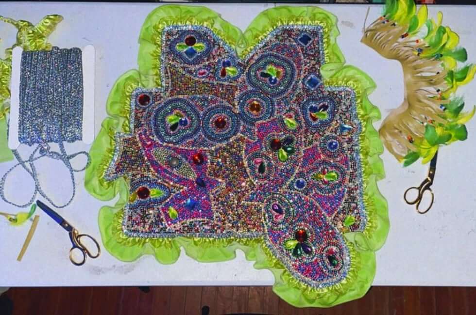 A patch in progress on a white table. The patch is covered in multi-colored rhinestones and bordered with silky lime green ruffles. The rhinestones create various geometric shapes with clear borders and additional gemstone embellishments inside. A pack of rhinestones lies on the left side of the patch, alongside gold scissors and scrap material. A semi-circle of green feathers lies to the right, also alongside a pair of gold scissors.
