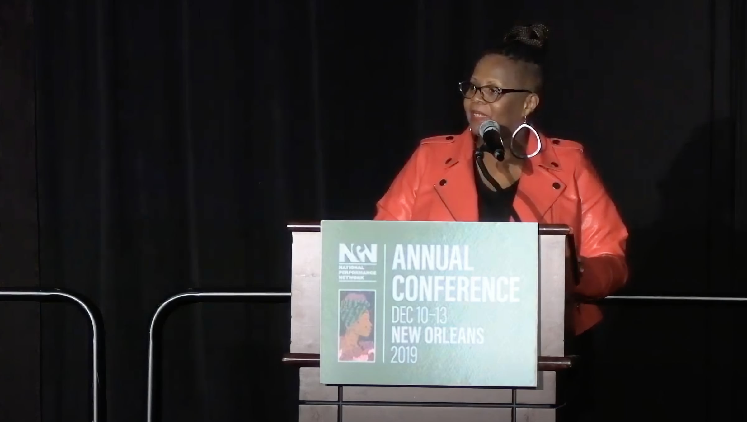 A woman wearing glasses and a bright orange jacket speaks at a podium on a dark stage, looking to the side as she addresses the audience. A microphone is positioned in front of her, and a large sign on the podium reads Annual Conference Dec 10 to 13 New Orleans 2019. The focused lighting highlights her presence and emphasizes the formal conference setting.