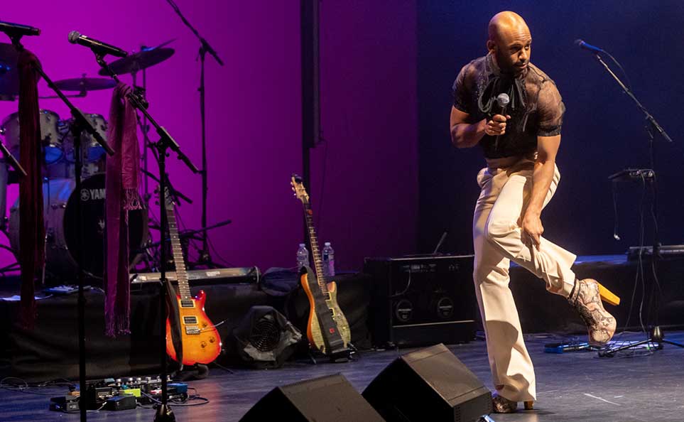 Joseph Hall, Co-Executive Director of Kelly Strayhorn Theater, on stage showing off his high-heel boots to the audience, lifting one foot with a grin while holding a microphone. He wears a sheer black top and cream-colored wide-leg pants. Guitars rest on stands near a drum kit behind him, lit by dramatic magenta and deep blue stage lighting.