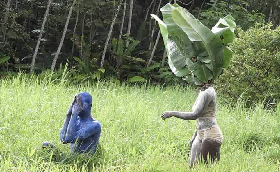 Two figures in a lush green field with tall grass and tropical forest behind them. One, covered entirely in blue body paint, sits crouching with hands pressed together near their face. The other, covered in white or gray body paint, stands with a large banana leaf draped over their head and shoulders, one arm extended. Both appear to be part of a land-based performance.