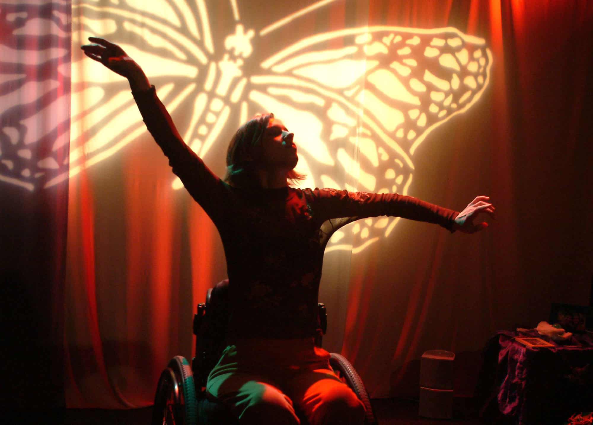 Person in a wheelchair poses with arms extended in a graceful dance position on a dimly lit stage. A large glowing butterfly projection fills the backdrop against red curtains, creating a dramatic and empowering performance scene. The warm orange and red lighting highlights the movement and artistry of inclusive dance.