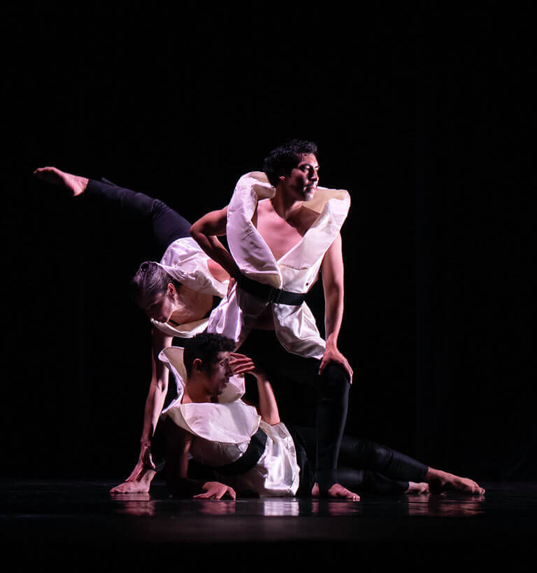 Three contemporary dancers in sculptural white costumes with wide collars and black belts perform an intricate, balanced pose on a dark stage. Their limbs interlock as one dancer supports another’s raised leg while the third crouches at the base, creating a striking triangular composition.