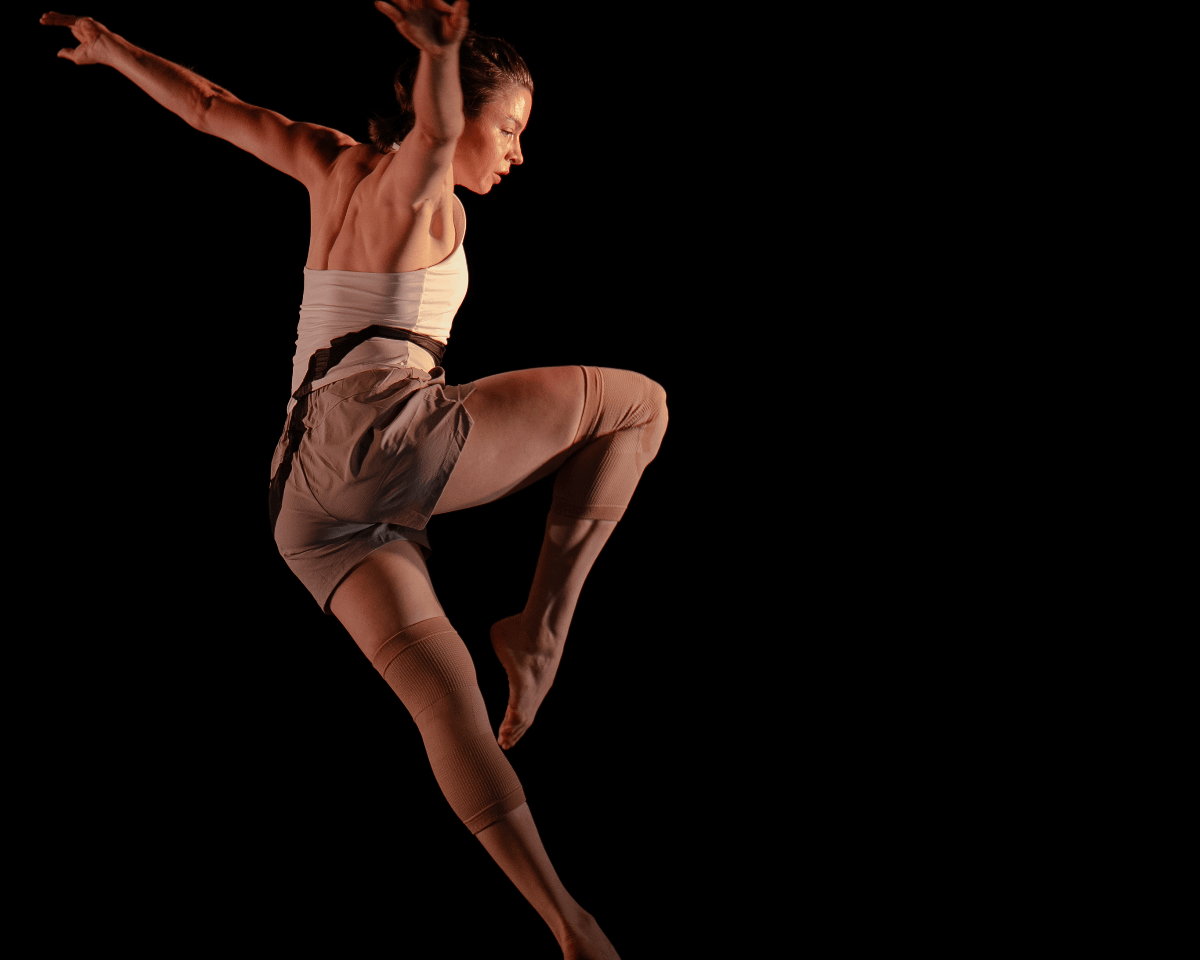 A contemporary dancer is captured mid-air in a dynamic leap, with arms extended and one knee lifted high. She wears a fitted white top, beige shorts, and thigh-high leg warmers, with stage lighting highlighting the muscles in her back against a black background.