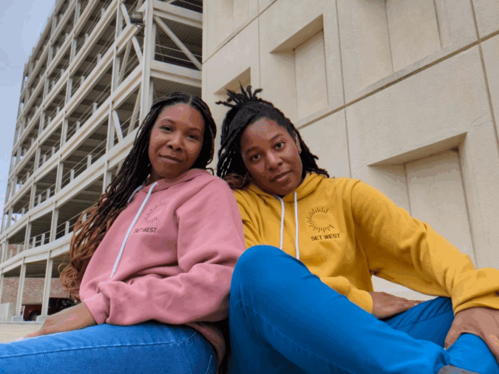 Two young Black women in jeans and sweatshirts sit outside and smile gently at the camera. On the left, Chasah West, wearing a muted pink sweatshirt, sits facing away from the camera with her back against her sister's shoulder and her face turned toward us. On the right, Charliese West, wearing a gold colored sweatshirt, sits cross-legged facingthe camera.