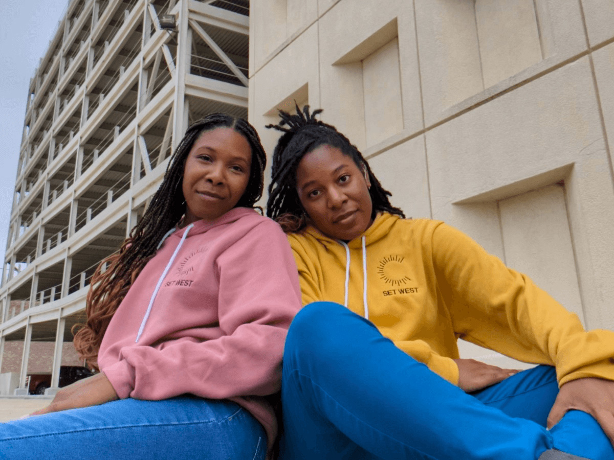 Two young Black women in jeans and sweatshirts sit outside and smile gently at the camera. On the left, Chasah West, wearing a muted pink sweatshirt, sits facing away from the camera with her back against her sister's shoulder and her face turned toward us. On the right, Charliese West, wearing a gold colored sweatshirt, sits cross-legged facingthe camera.