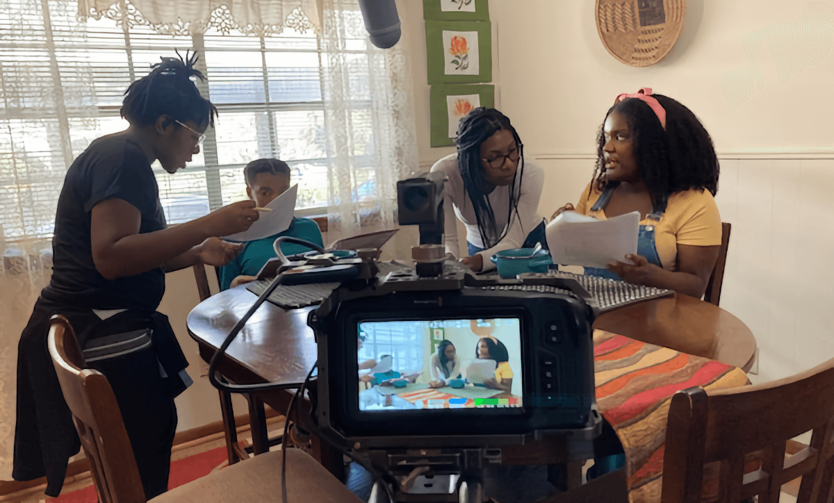 A behind-the-scenes shot of The West Sisters co-directing a scene from their short film "Héritage." Four young Black women are gathered around a large table in a brightly lit dining room. Behind them on the left is a large, wide window covered in sheer lace curtains that let in plenty of outside light. On the left side of the table, standing in profile and holding pages of a screenplay, Charliese West speaks to one of the actresses, who is sitting at the table immediately to her right. Chasah West is in the center of the image, standing but bent over with her forearms resting on the table, as she consults with the second actress, who is sitting to her right and holding pages of the screenplay. Above them is the end of a boom microphone, and in the extreme center foreground is a DSLR camera with the same composition in the photo framed in miniature on the digital screen.